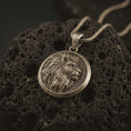 Load image into Gallery viewer, Bronze lion medallion pendant necklace with an oxidized finish resting on textured volcanic rock, captured in a close-up premium studio composition.
