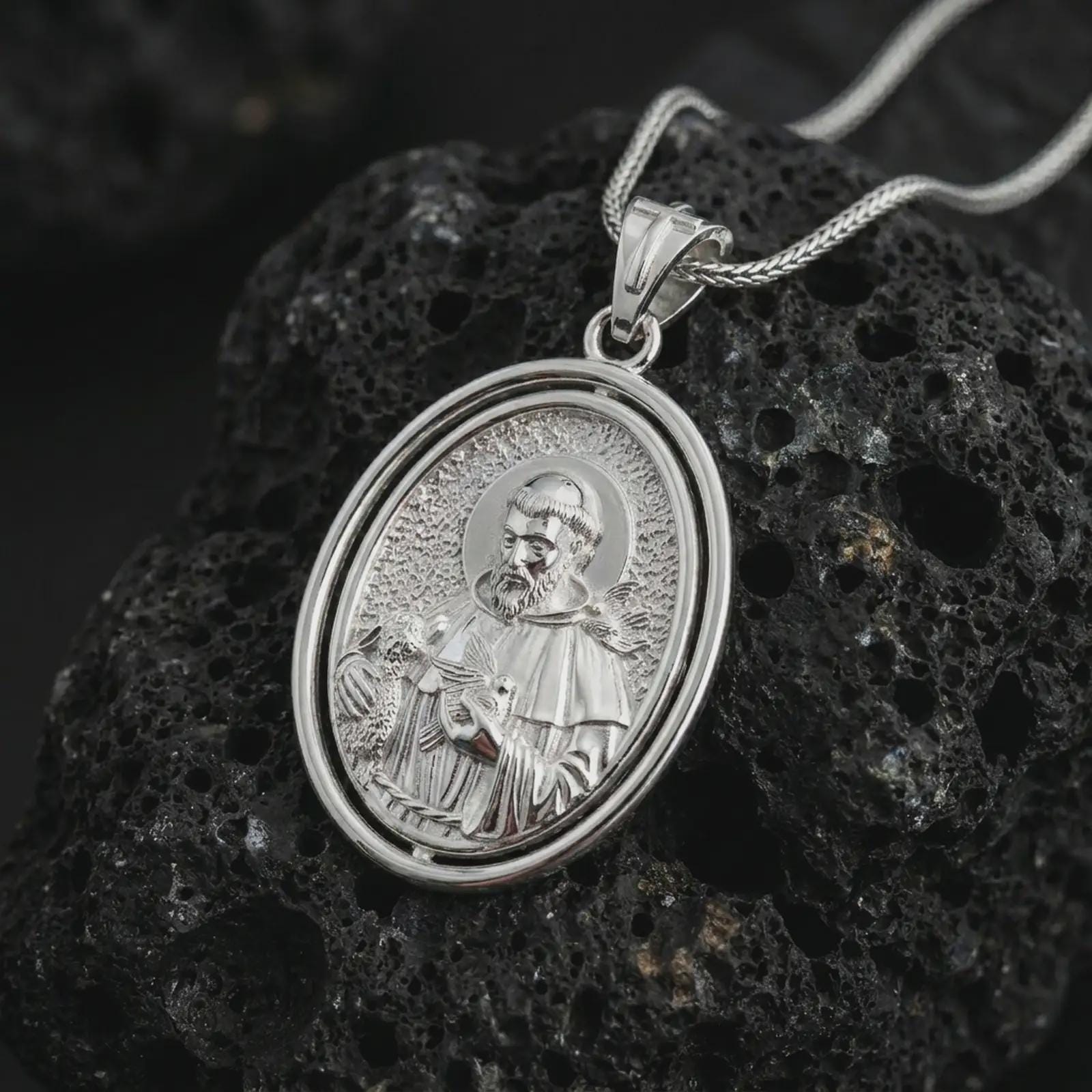 Polished silver Saint Francis pendant necklace on textured volcanic rock, centered macro jewelry photo with detailed Catholic medal relief and bright silver highlights.