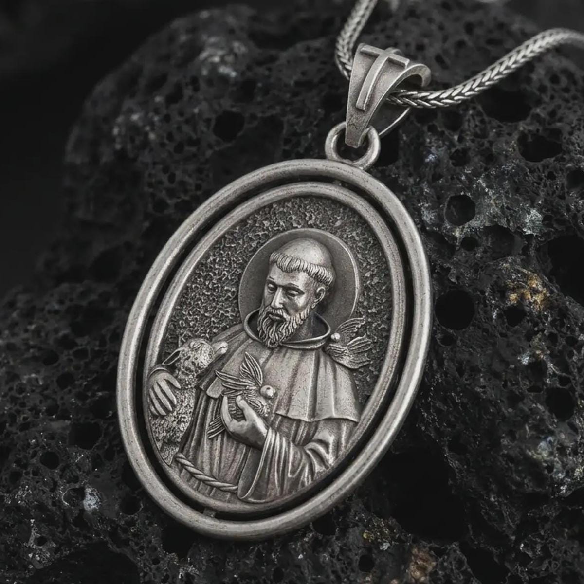 Oxidized Saint Francis pendant necklace on textured volcanic rock, close-up studio shot highlighting the saint relief, lamb, birds, and antique silver contrast.