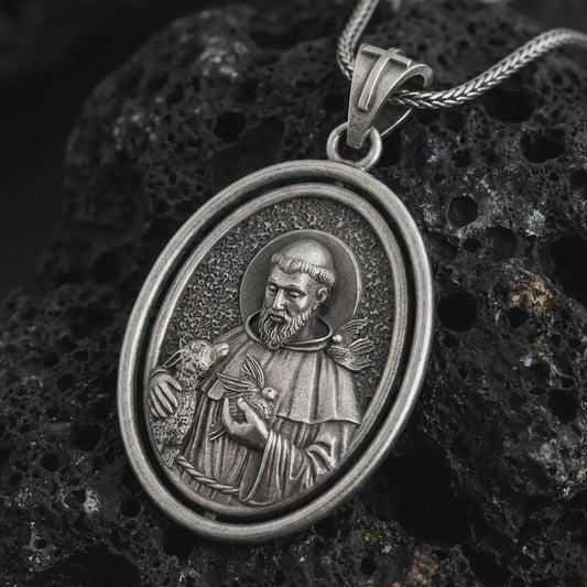 Oxidized Saint Francis pendant necklace on textured volcanic rock, close-up studio shot highlighting the saint relief, lamb, birds, and antique silver contrast.