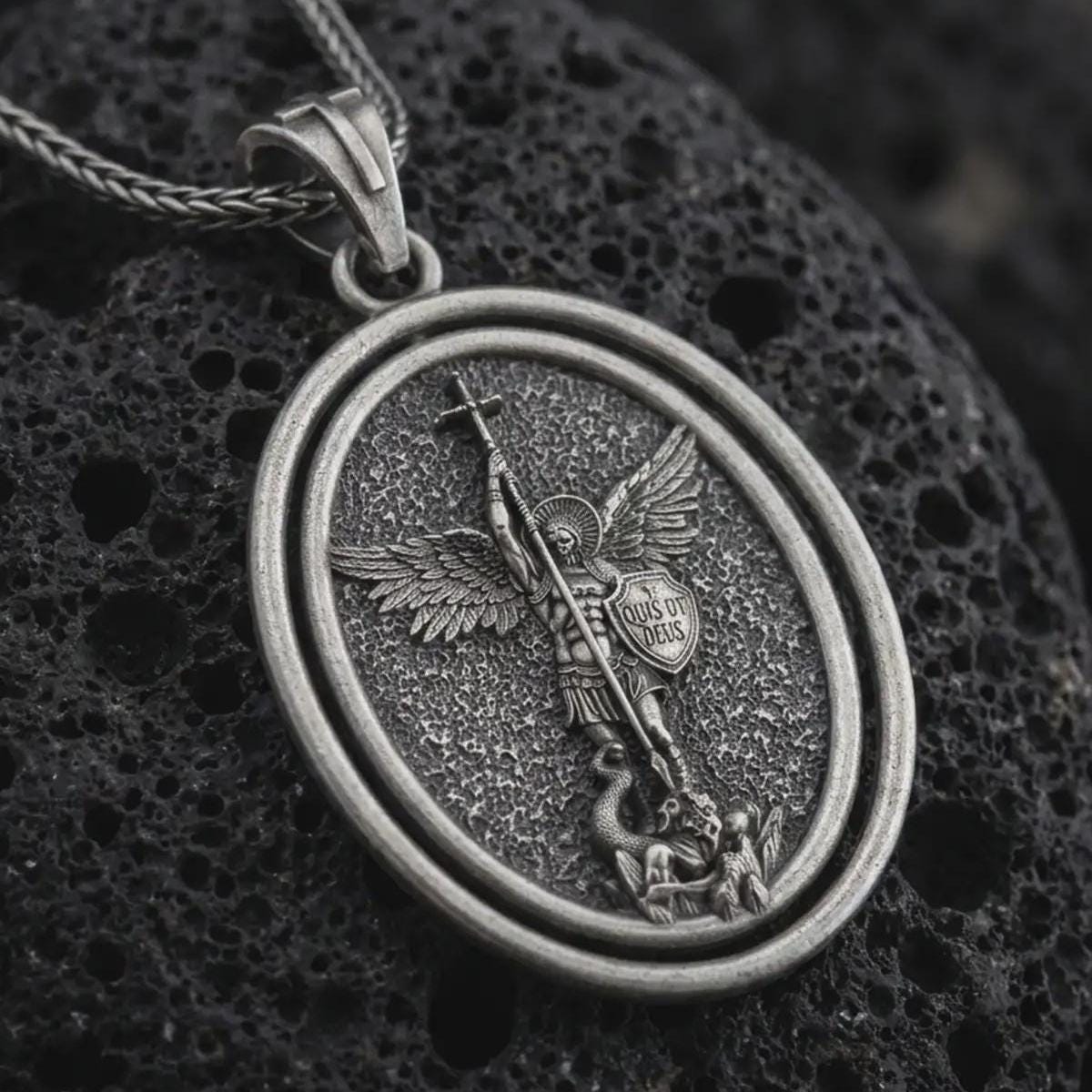 Oxidized Saint Michael spinning pendant necklace on textured volcanic rock, close-up studio shot highlighting the archangel wings, sword, shield, fallen demon, and antique silver contrast.
