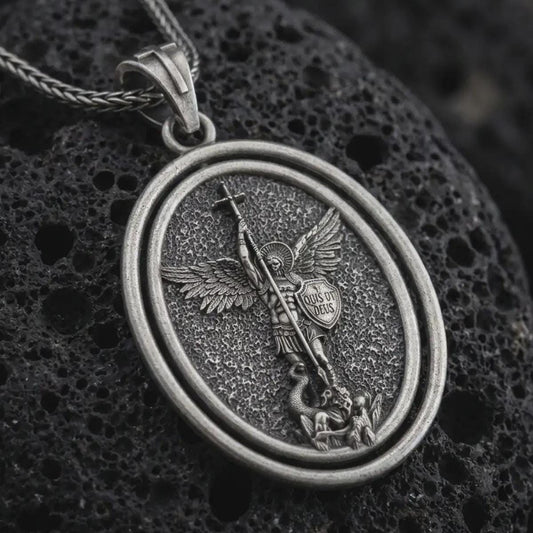 Oxidized Saint Michael spinning pendant necklace on textured volcanic rock, close-up studio shot highlighting the archangel wings, sword, shield, fallen demon, and antique silver contrast.