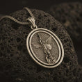 Load image into Gallery viewer, Bronze Saint Michael spinning pendant necklace on textured volcanic rock, close-up studio shot highlighting the archangel wings, sword, shield, fallen demon, and antique silver contrast.
