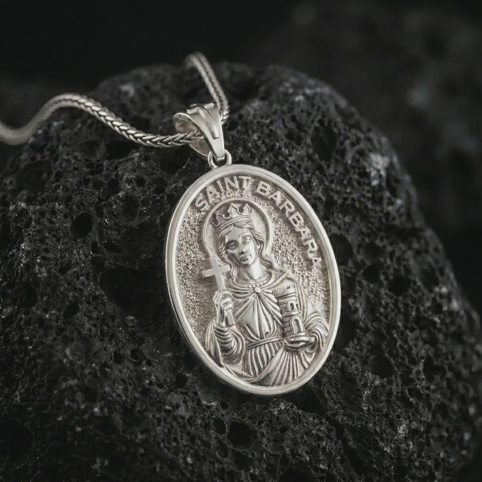 Polished silver Saint Barbara pendant necklace on textured volcanic rock, centered macro jewelry photo with detailed Catholic medal engraving and bright silver highlights.