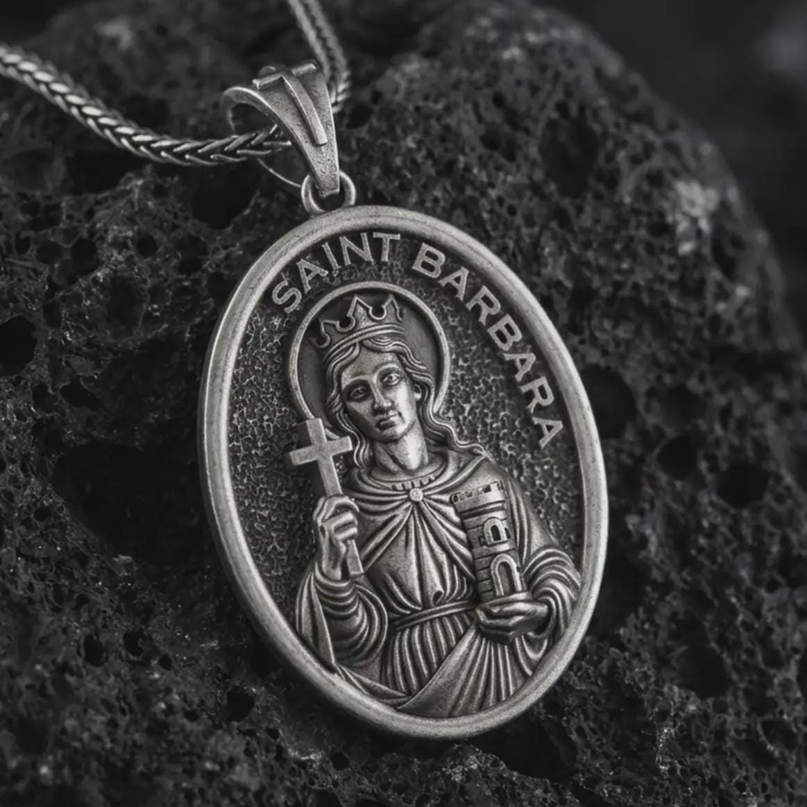 Oxidized Saint Barbara pendant necklace on textured volcanic rock, close-up studio shot highlighting the crowned saint relief, cross, tower detail, and antique silver contrast.