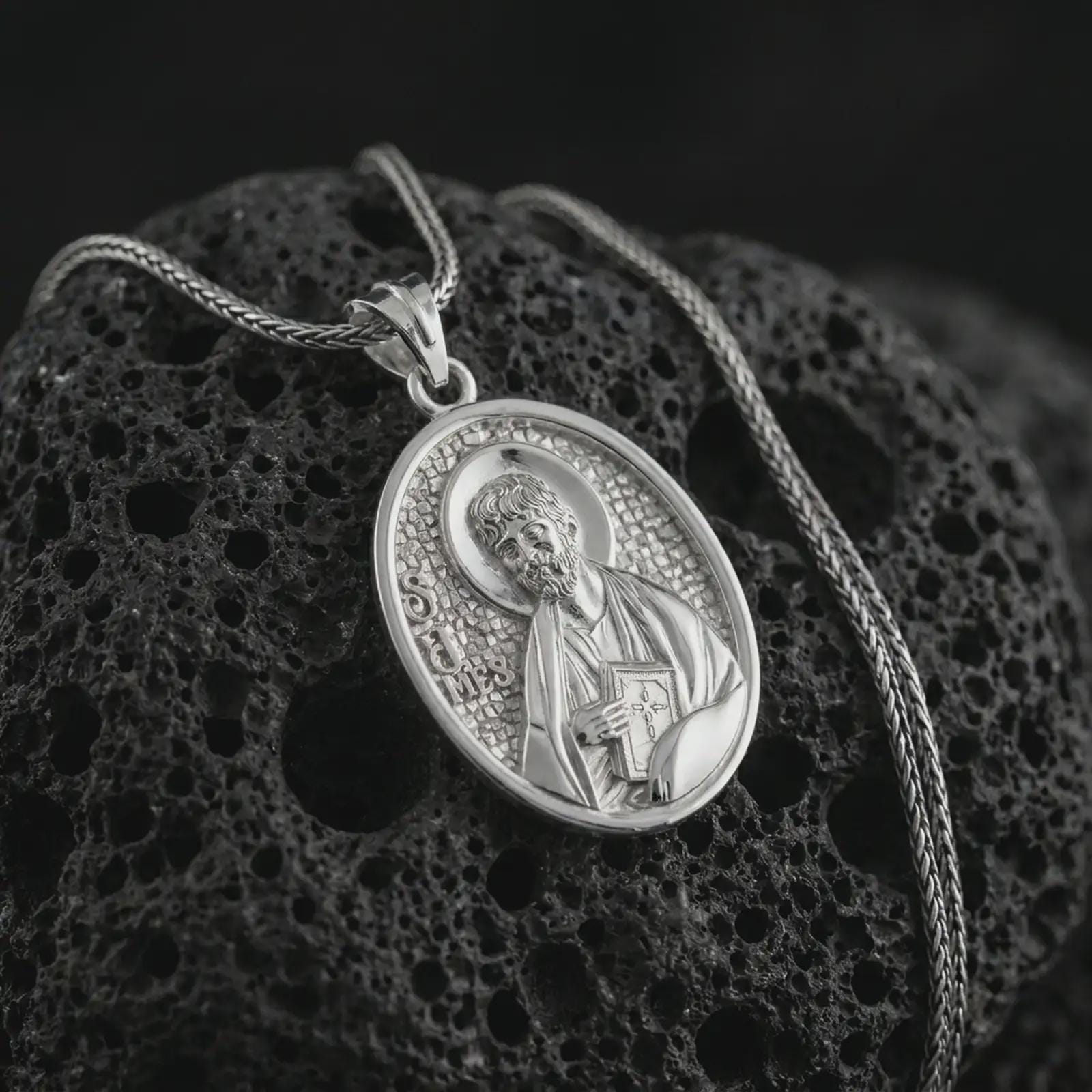 Polished silver Saint James the Apostle pendant necklace on textured volcanic rock, centered macro jewelry photo with detailed religious medal engraving and bright silver highlights.