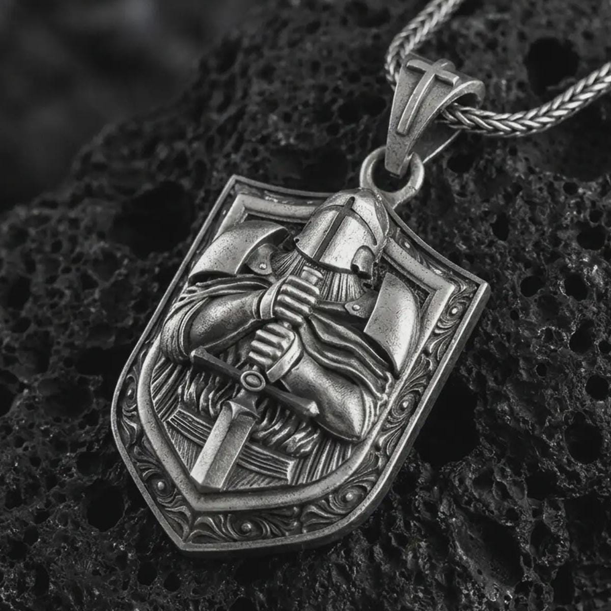 Oxidized knight shield pendant necklace on textured volcanic rock, close-up studio shot highlighting the armored crusader figure, sword, shield shape, and antique silver contrast.