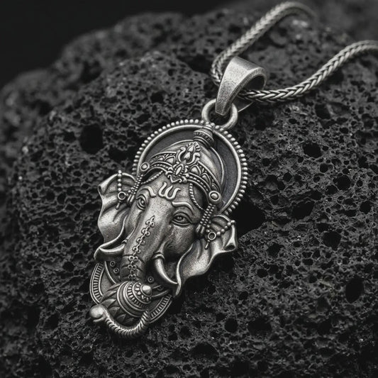 Oxidized Ganesha pendant necklace on textured volcanic rock, close-up studio shot highlighting the elephant god face, ornate crown, tusks, and antique silver contrast.