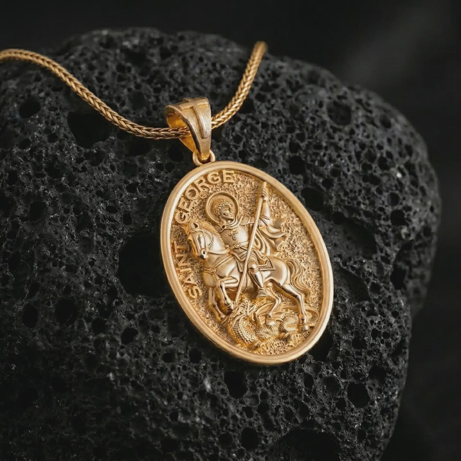 Gold plated Saint George pendant necklace on textured volcanic rock, premium close-up studio composition emphasizing the horse, spear, and defeated dragon symbolism.