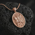Load image into Gallery viewer, Rose gold plated Saint George pendant necklace on textured volcanic rock, detailed macro product shot with soft warm reflections and heroic saint design.
