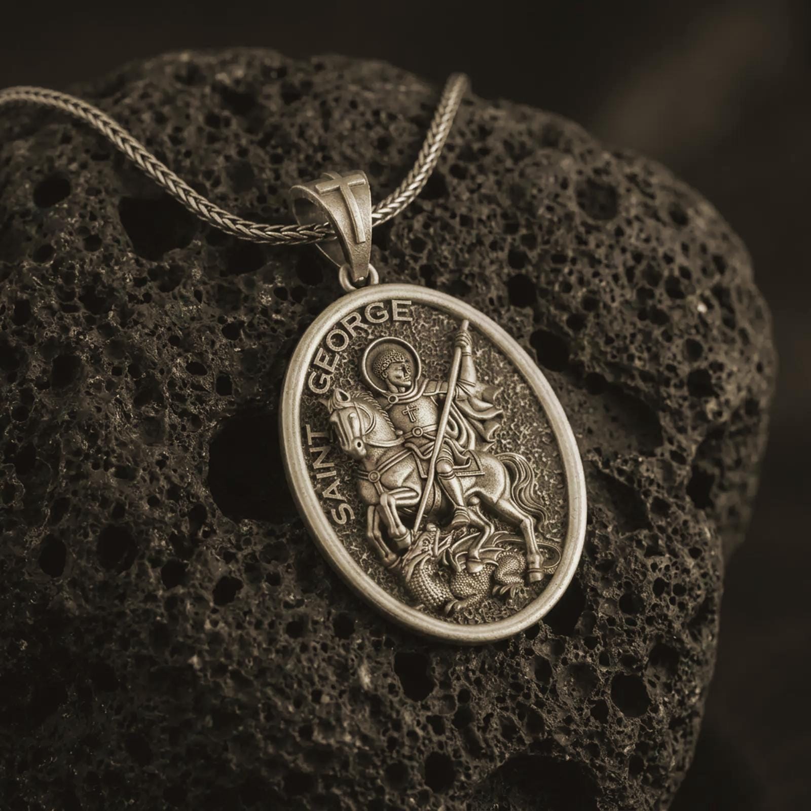 Bronze Saint George pendant necklace on textured volcanic rock, close-up studio shot highlighting the mounted saint, spear, dragon detail, and antique silver contrast.