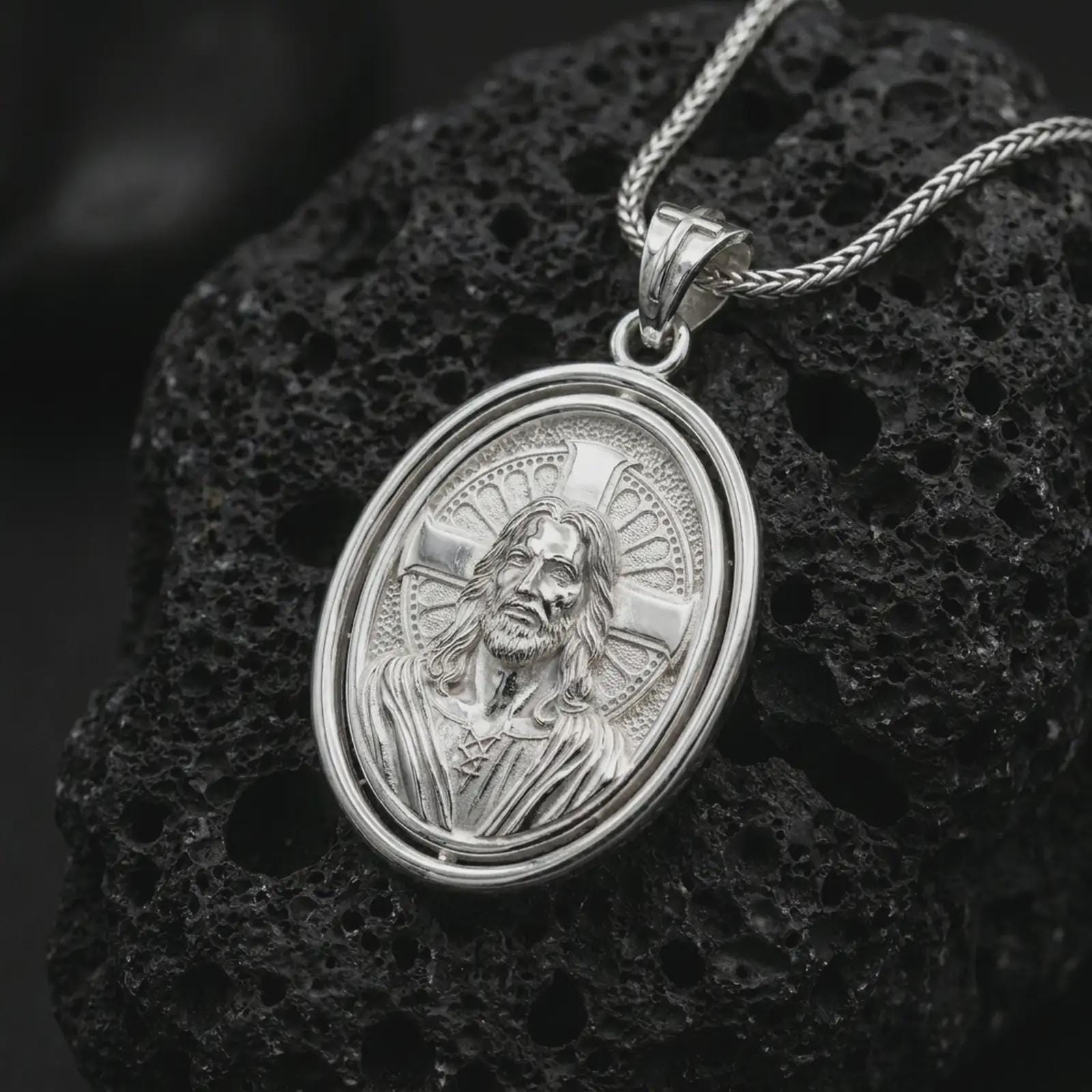 Polished silver spinning Jesus Christ pendant necklace on textured volcanic rock, centered macro jewelry photo with detailed Christian portrait relief and bright silver highlights.