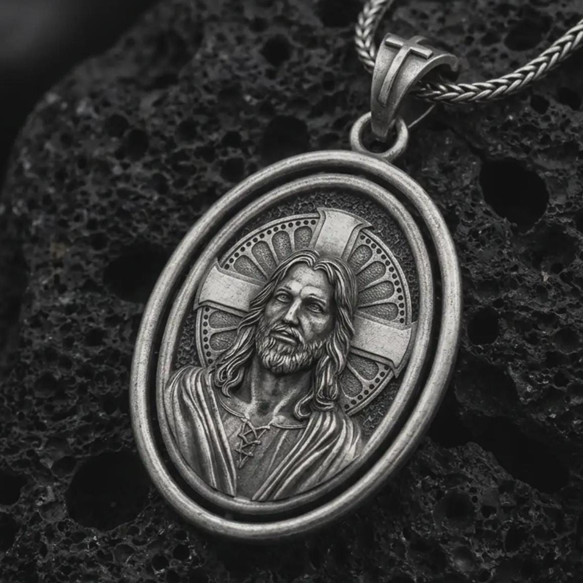 Oxidized spinning Jesus Christ pendant necklace on textured volcanic rock, close-up studio shot highlighting the Jesus portrait, cross halo, oval frame, and antique silver contrast.
