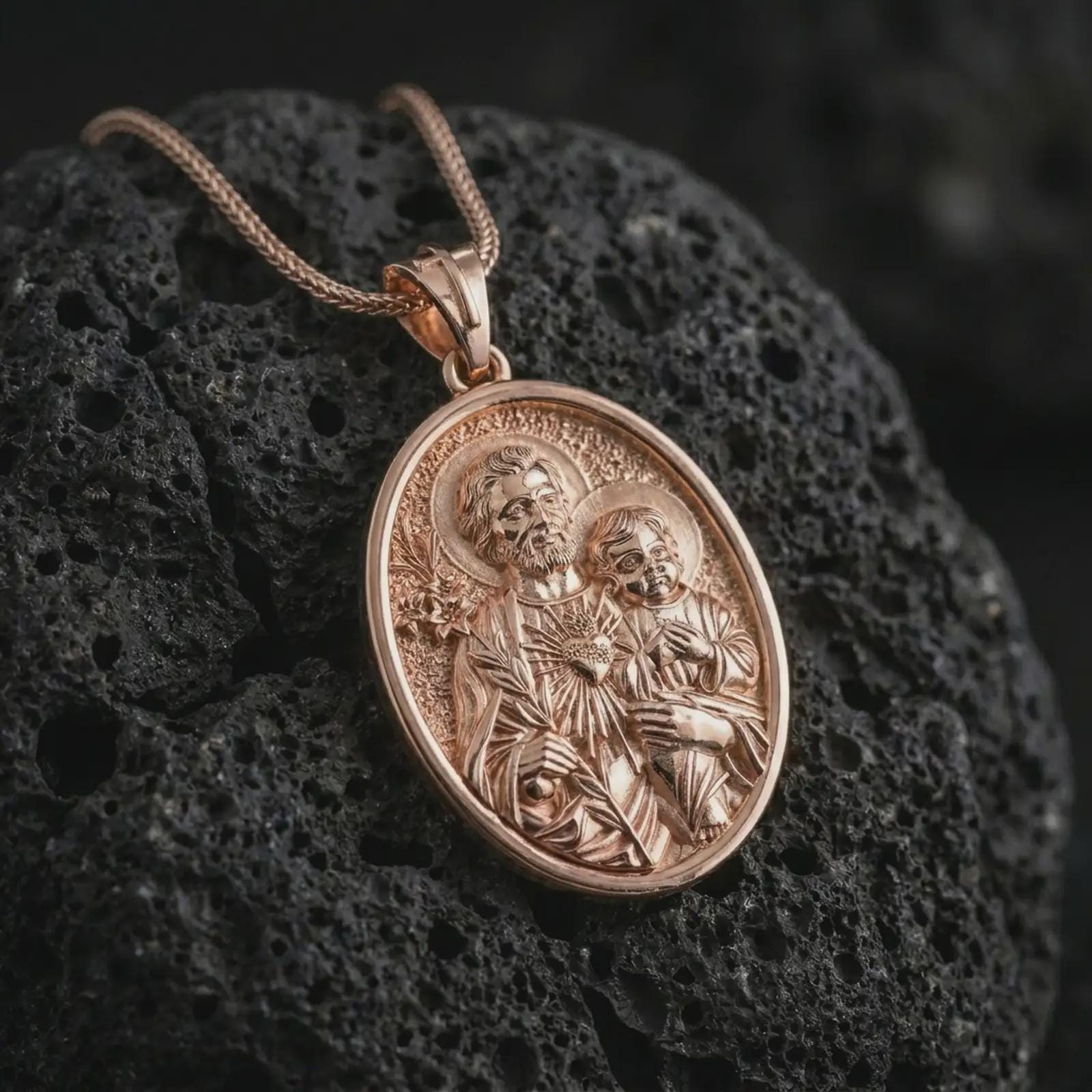 Rose gold plated Saint Joseph and Jesus pendant necklace on textured volcanic rock, detailed macro product shot with soft warm reflections and devotional father-and-child design.