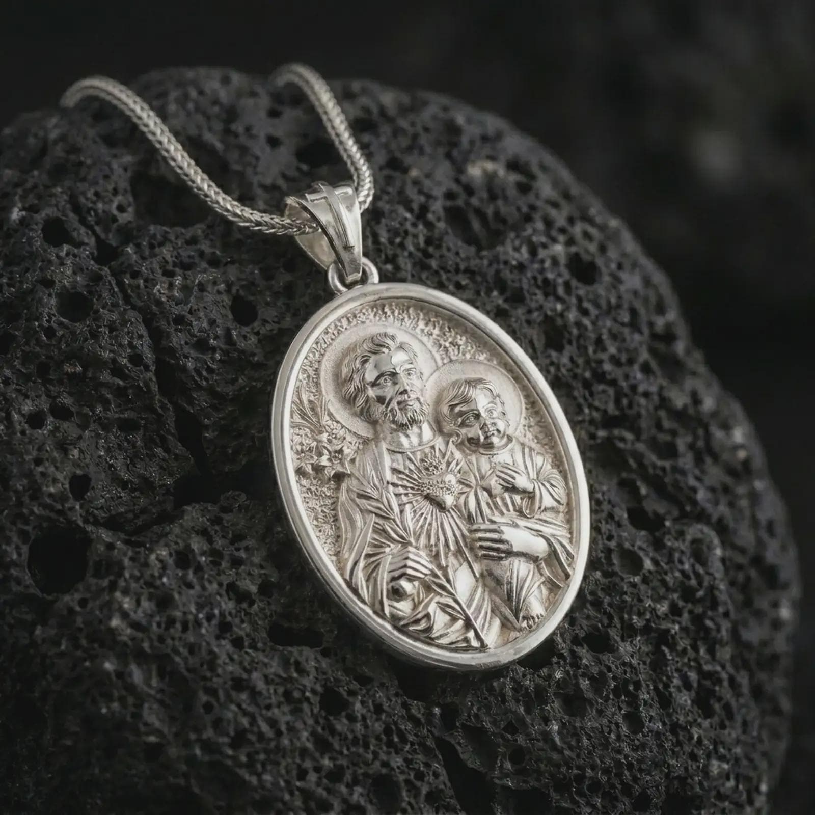 Polished silver Saint Joseph and Jesus pendant necklace on textured volcanic rock, centered macro jewelry photo with detailed Catholic medal engraving and bright silver highlights.
