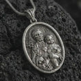 Load image into Gallery viewer, Oxidized Saint Joseph and Jesus pendant necklace on textured volcanic rock, close-up studio shot highlighting the father and child relief, lily, sacred heart, and antique silver contrast.
