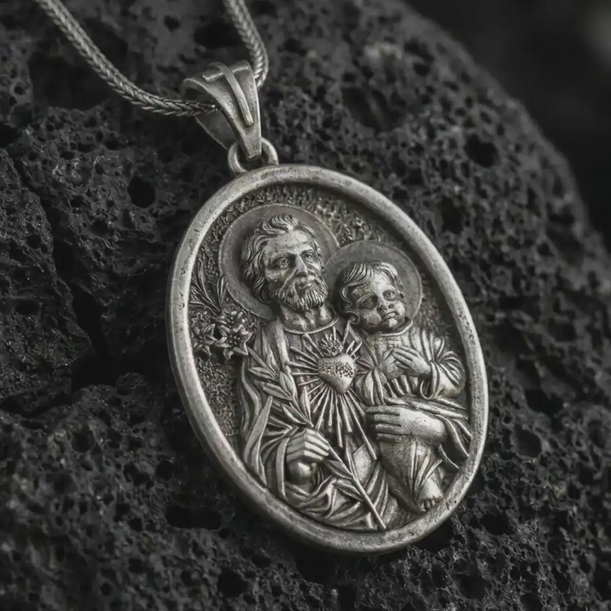 Oxidized Saint Joseph and Jesus pendant necklace on textured volcanic rock, close-up studio shot highlighting the father and child relief, lily, sacred heart, and antique silver contrast.