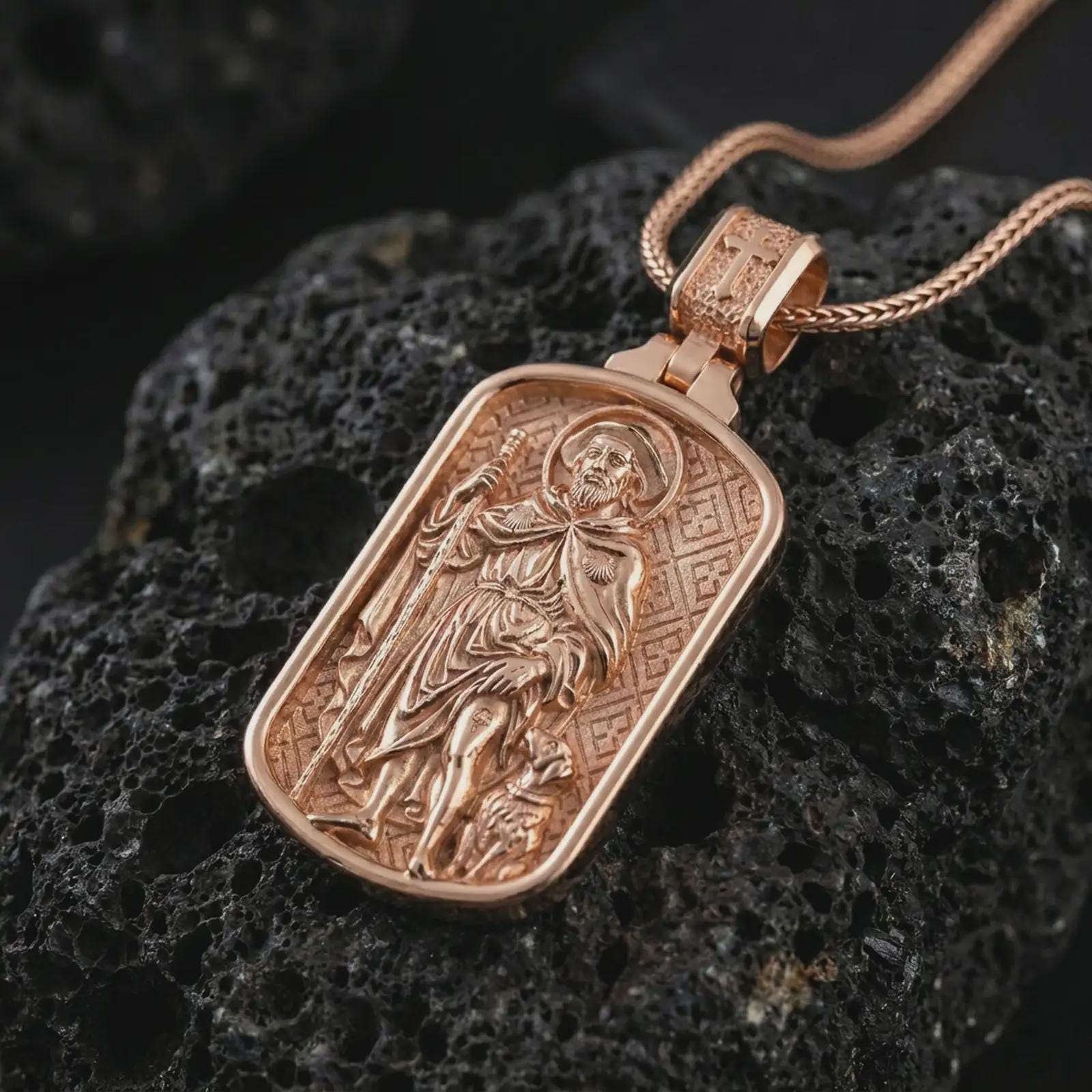 Rose gold plated Saint Roch dog tag pendant necklace on textured volcanic rock, detailed macro product shot with soft warm reflections and devotional Saint Roch design.