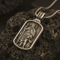 Load image into Gallery viewer, Bronze Saint Roch dog tag pendant necklace on textured volcanic rock, close-up studio shot highlighting the pilgrim saint relief, staff, dog detail, and antique silver contrast.
