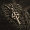 Load image into Gallery viewer, Bronze Celtic cross pendant necklace with an oxidized finish displayed on black volcanic stone, captured in a close-up studio image highlighting the bold cross form and circular halo detail.
