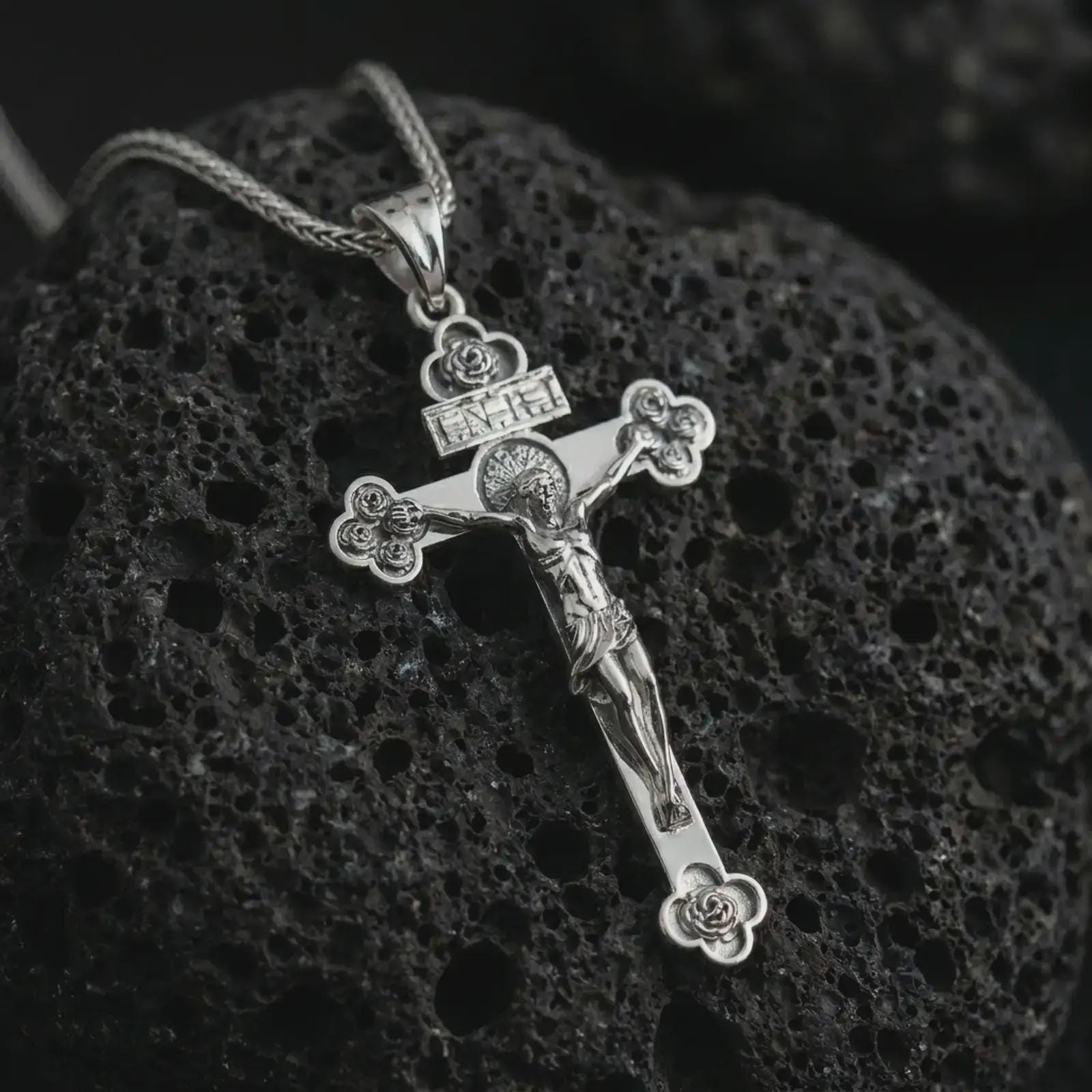 Polished silver crucifix pendant necklace resting on textured volcanic rock, shown in a premium macro product shot that emphasizes the sacred cross design and refined religious detailing.