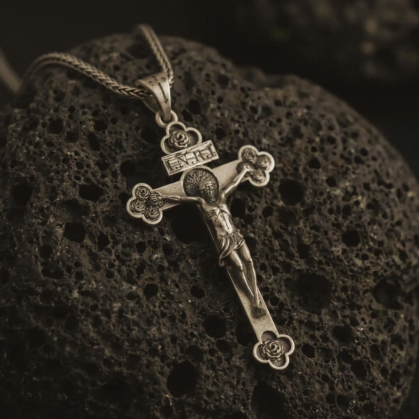 Bronze crucifix pendant necklace with an oxidized finish displayed on black volcanic stone, captured in a close-up studio image highlighting the detailed Jesus figure and floral cross tips.