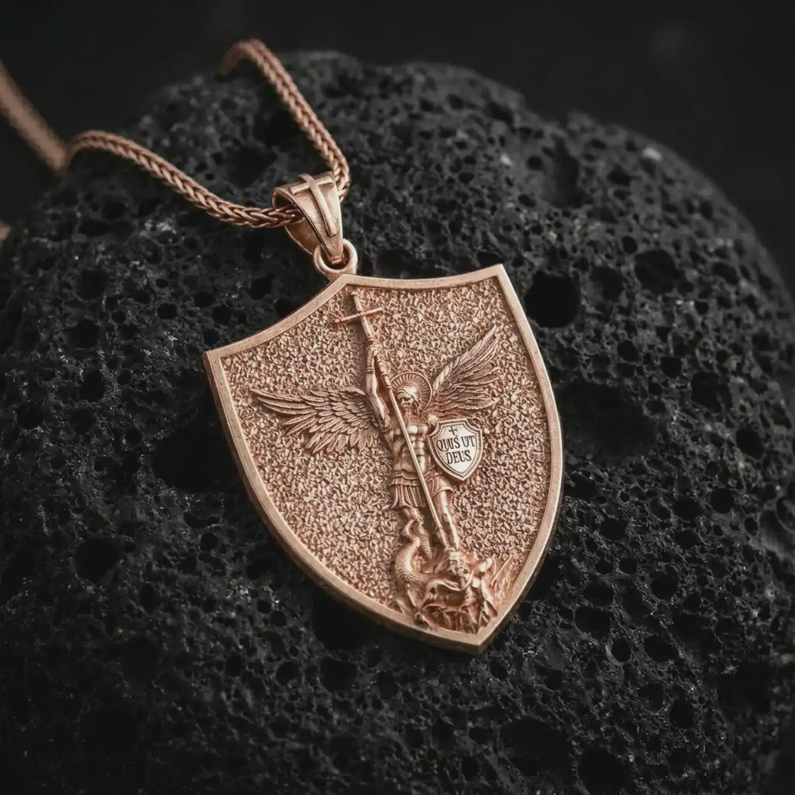 Rose gold plated Saint Michael pendant necklace photographed on porous volcanic stone, emphasizing the shield silhouette, sacred angel figure, and refined religious craftsmanship.