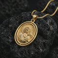 Load image into Gallery viewer, Gold plated Madonna and Child pendant necklace photographed on textured black volcanic stone, emphasizing the haloed Virgin Mary and Christ child in a refined Catholic pendant composition.
