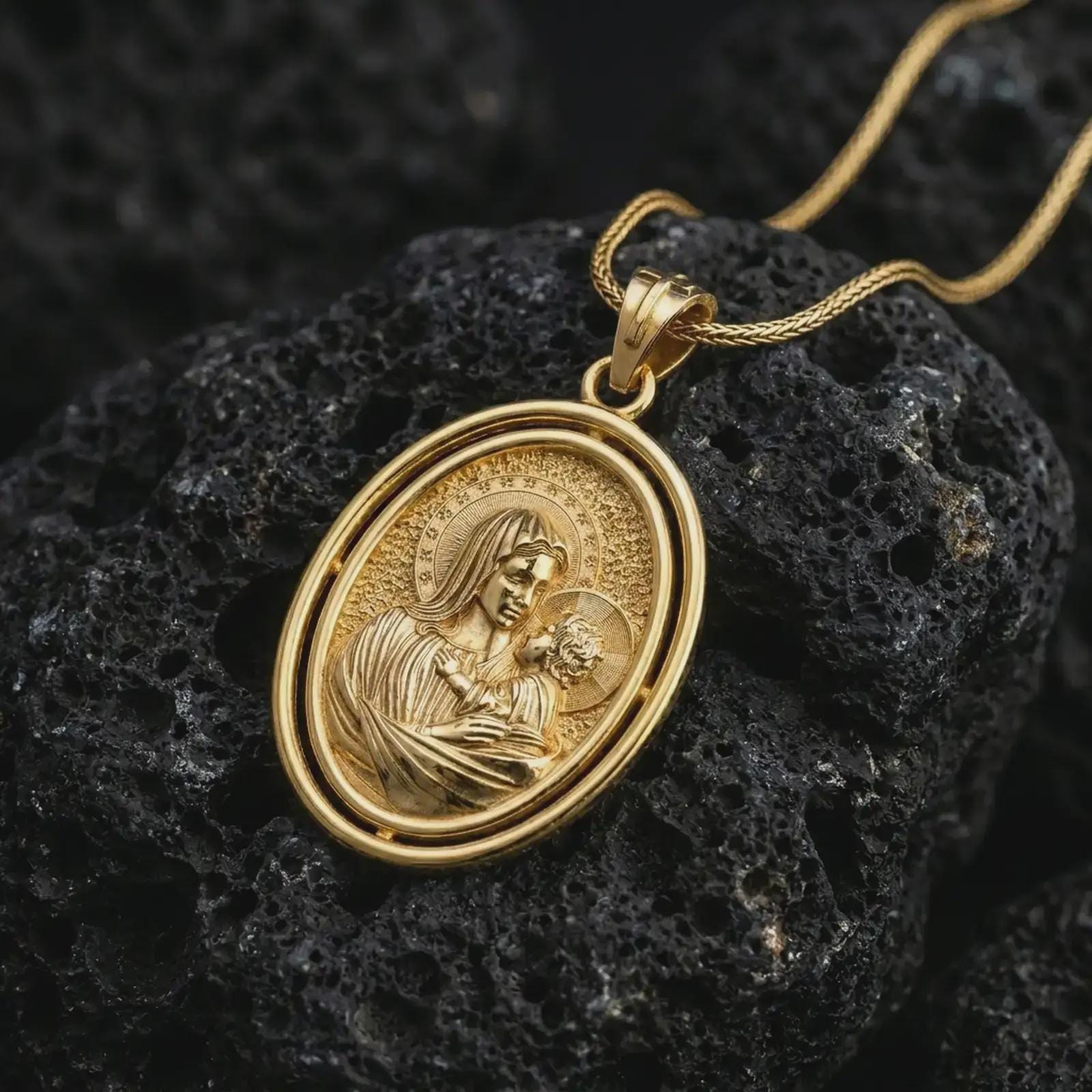 Gold plated Madonna and Child pendant necklace photographed on textured black volcanic stone, emphasizing the haloed Virgin Mary and Christ child in a refined Catholic pendant composition.