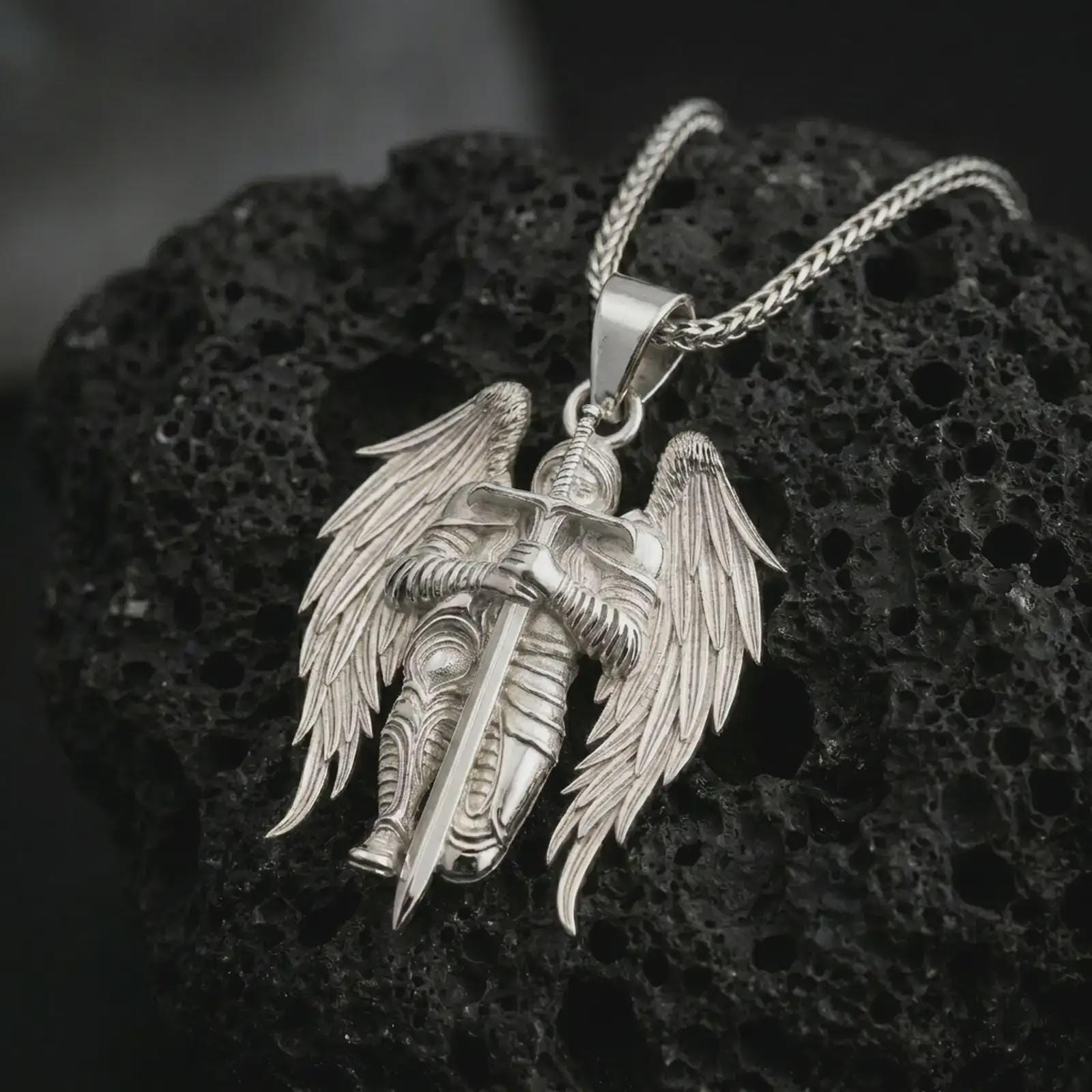 Polished silver angel warrior pendant necklace resting on dark volcanic rock, highlighting the armored celestial figure with spread wings and a strong protective stance.