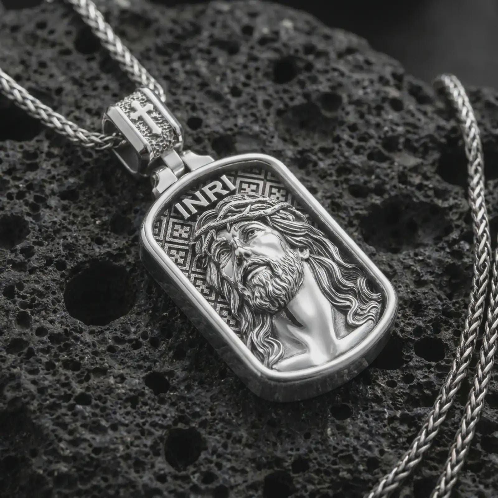 Polished silver INRI Jesus pendant necklace on textured volcanic rock, centered macro jewelry photo with detailed Christian portrait relief and bright silver highlights.