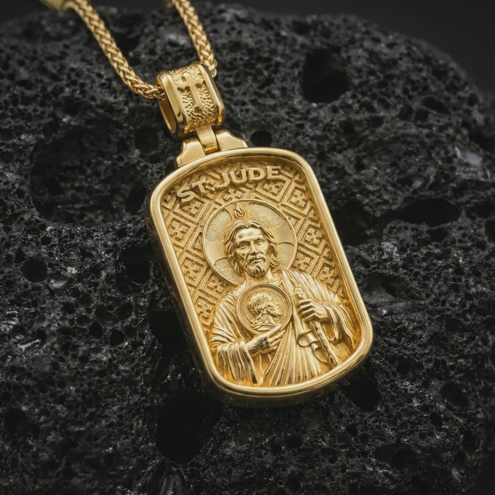 Gold plated Saint Jude dog tag pendant necklace on textured volcanic rock, premium close-up studio composition emphasizing the saint figure, halo, and sacred medallion symbolism.