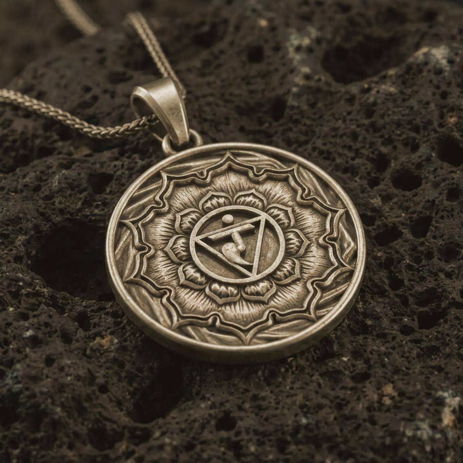 Bronze Manipura chakra pendant necklace on textured volcanic rock, close-up studio shot highlighting the solar plexus symbol, lotus petals, geometric center, and antique silver contrast.