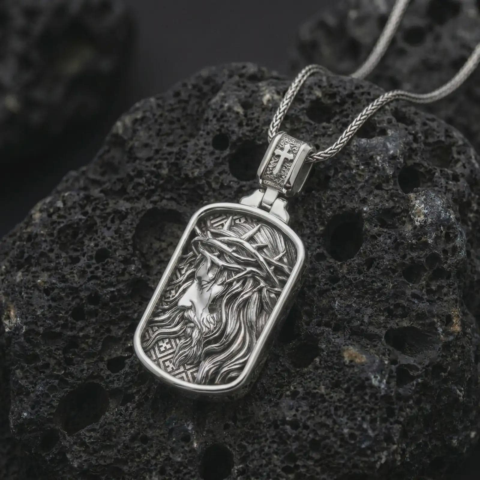 Polished silver crucified Jesus dog tag pendant necklace on textured volcanic rock, centered macro jewelry photo with detailed crown of thorns portrait and bright silver highlights.