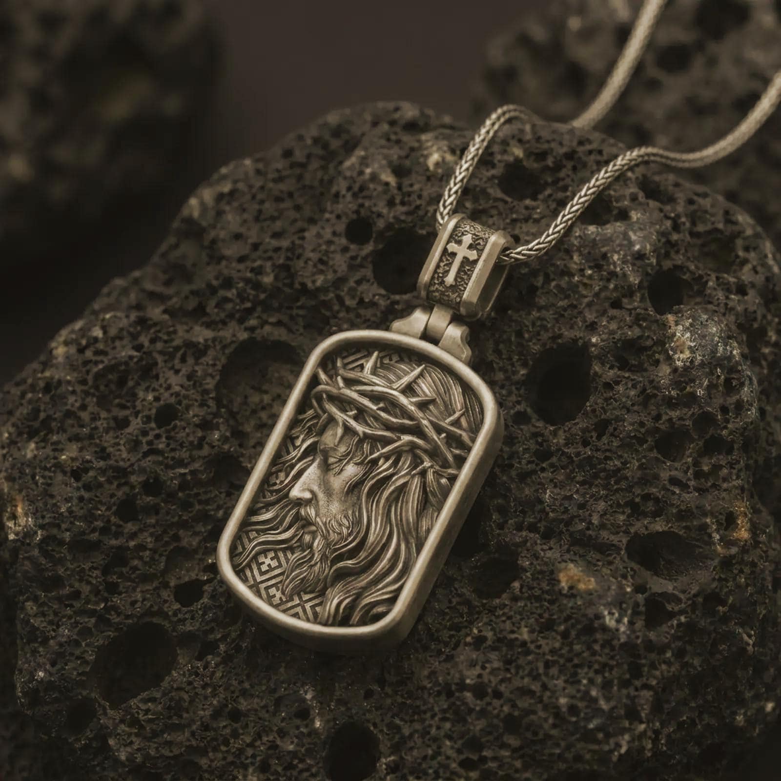 Bronze crucified Jesus dog tag pendant necklace on textured volcanic rock, close-up studio shot highlighting the side-profile Christ relief, crown of thorns, flowing hair, and antique silver contrast.