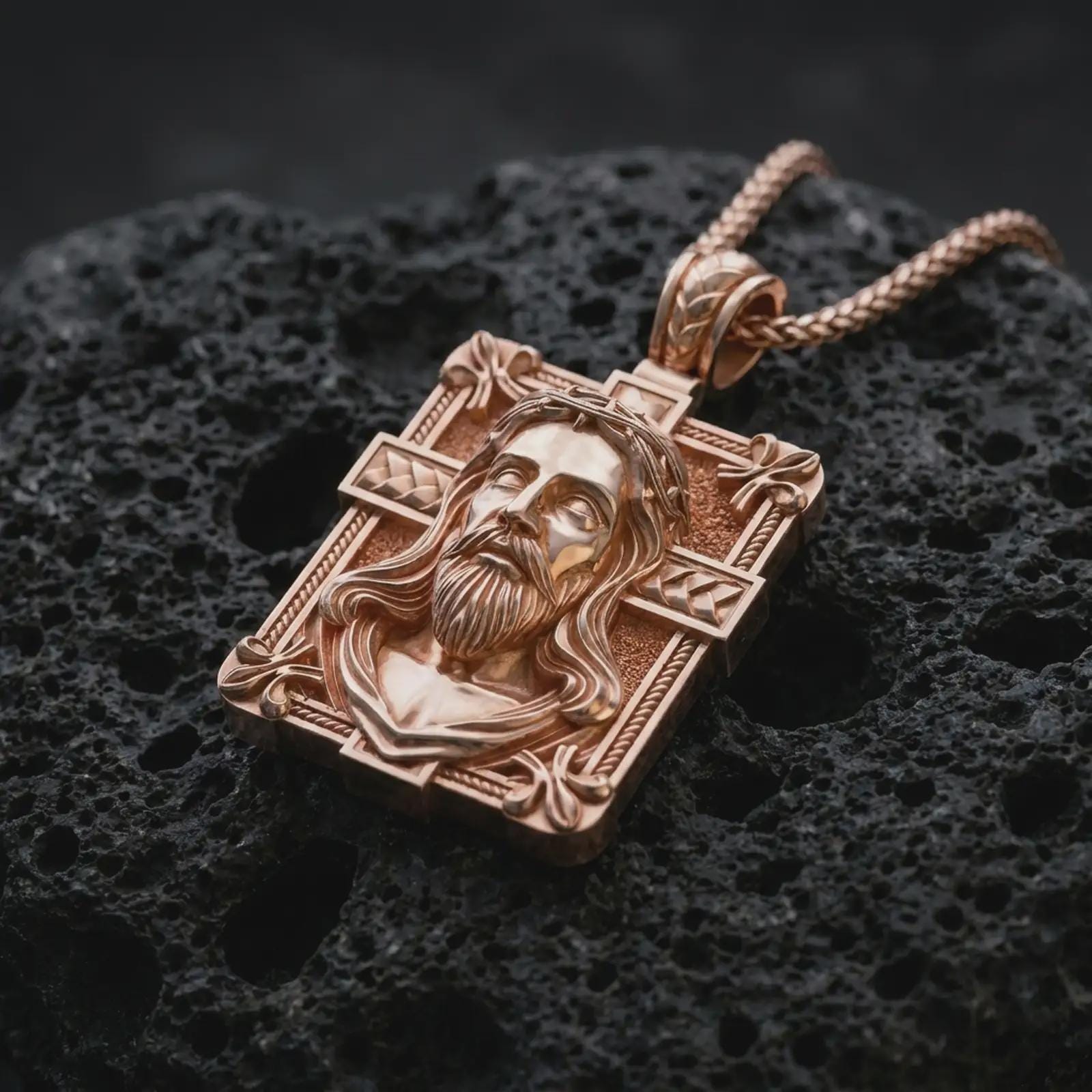 A Jesus crucifix pendant in a rose gold plated finish resting on porous volcanic rock in a premium macro studio shot.