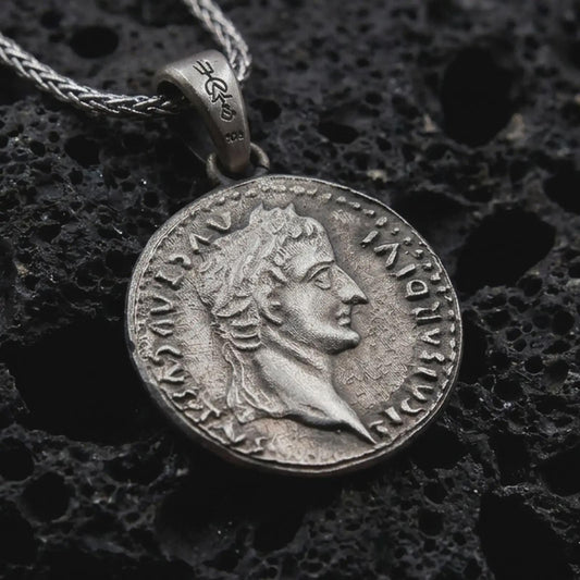 Oxidized Tiberius coin pendant necklace resting on black volcanic stone, featuring a Roman emperor profile and antique coin-style inscription in a detailed macro product photograph.