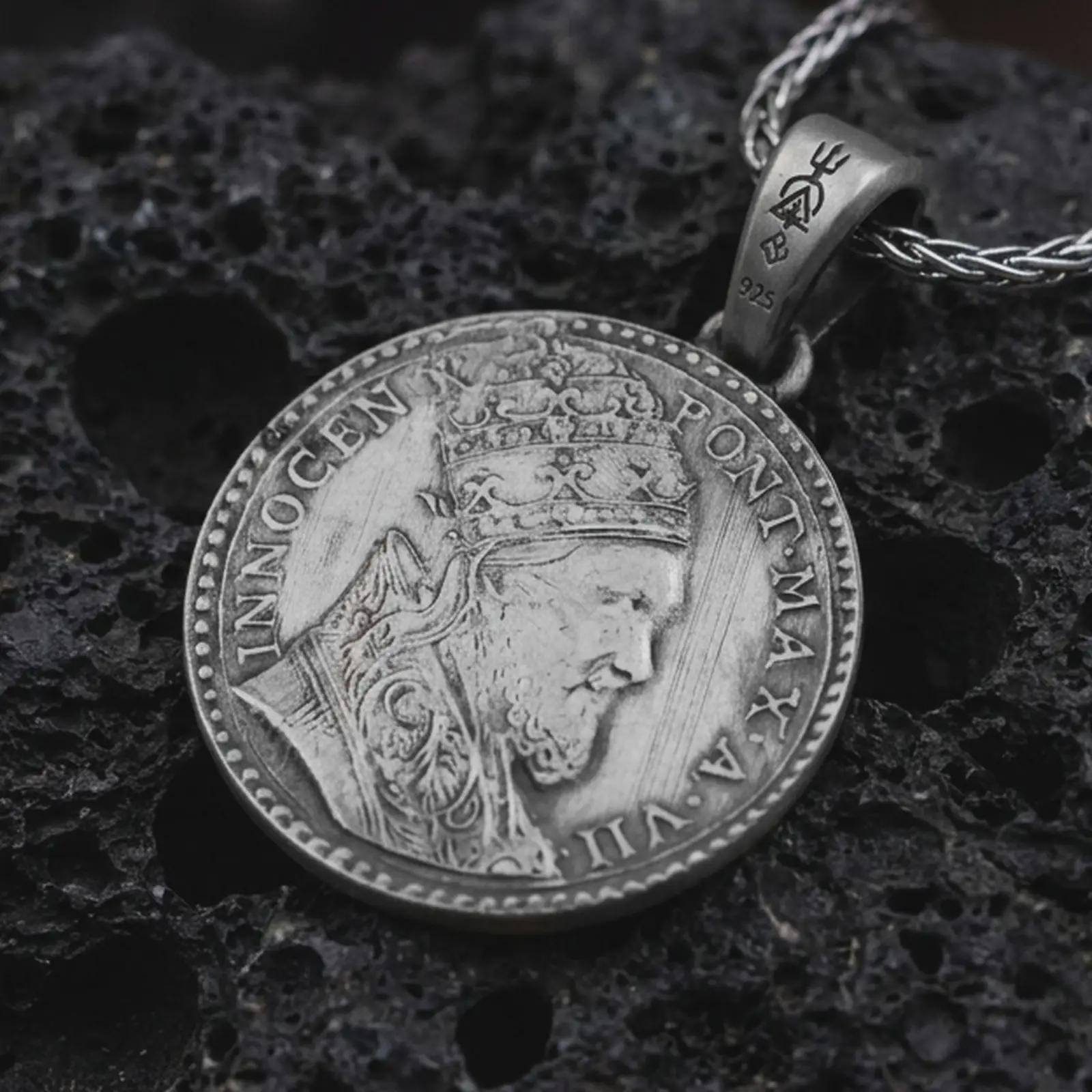 Oxidized Pope Innocent coin pendant necklace resting on black volcanic stone, featuring a papal portrait with Latin inscription in a detailed antique coin-style macro photograph.