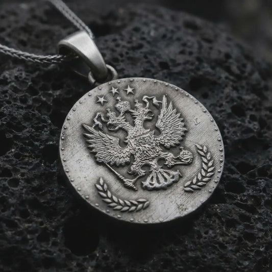 Oxidized heraldic eagle coin pendant necklace resting on black volcanic stone, featuring a round medallion with a crowned double-headed eagle, stars, and laurel branches in a detailed macro product photograph.