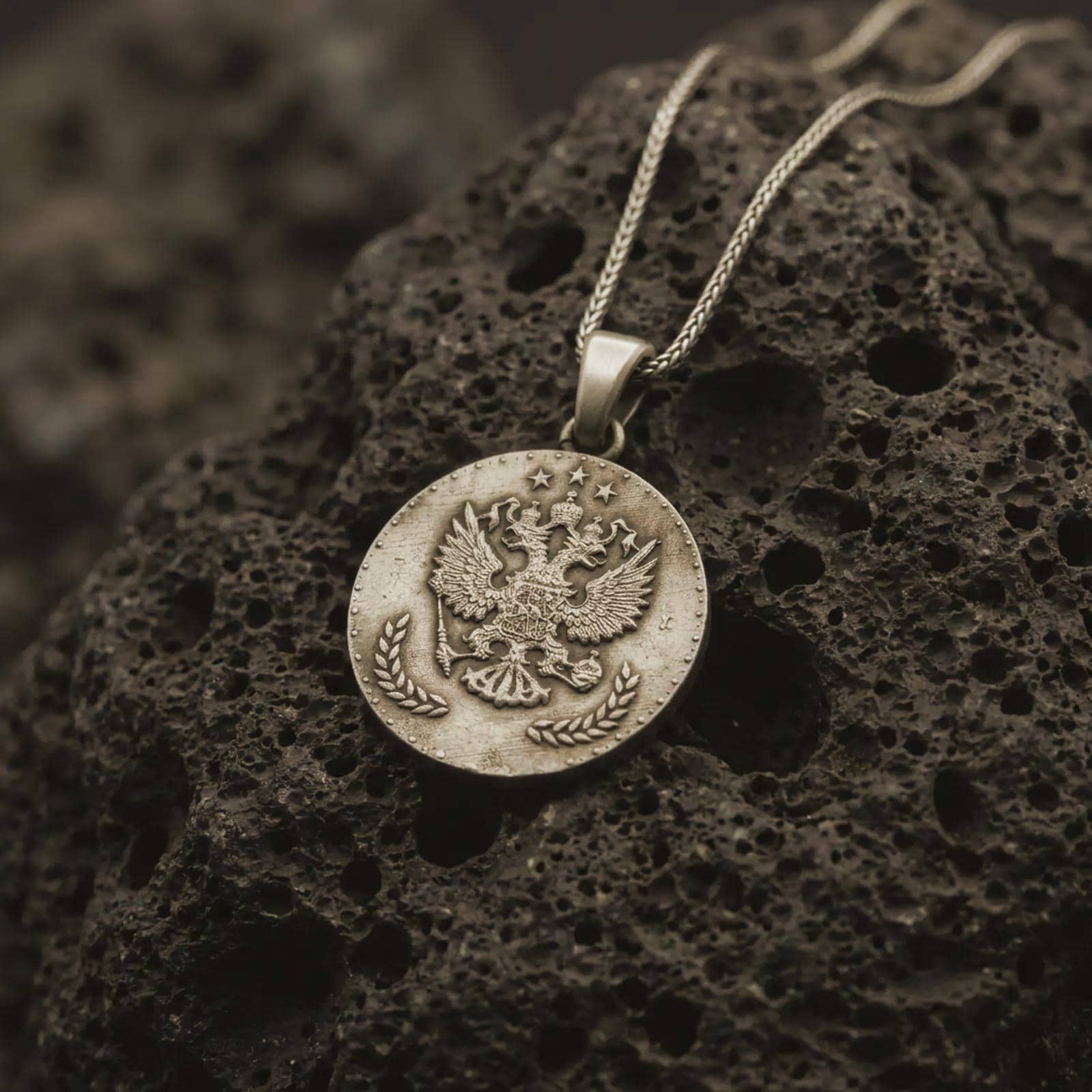 Bronze heraldic eagle coin pendant necklace resting on black volcanic stone, featuring a round medallion with a crowned double-headed eagle, stars, and laurel branches in a detailed macro product photograph.