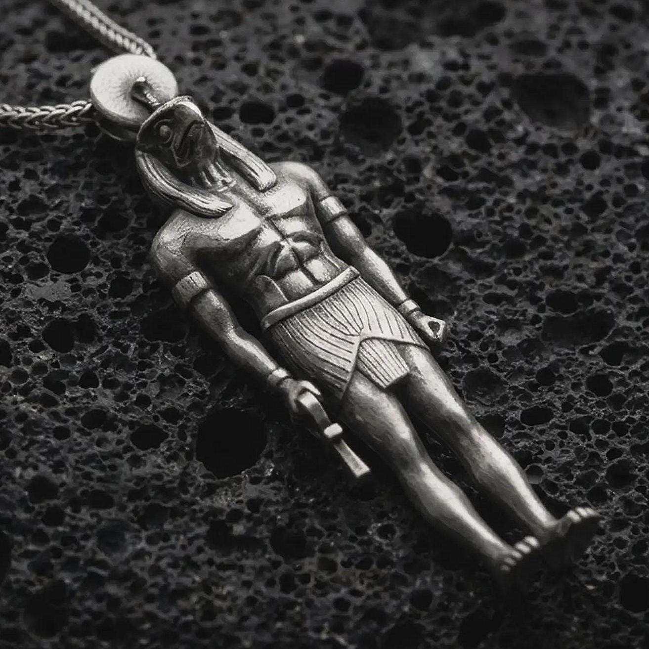 Oxidized Ra amulet pendant necklace resting on black volcanic stone, featuring an ancient Egyptian deity figure with a falcon-like head, sun disk, and strong sculpted body in a detailed macro product photograph.