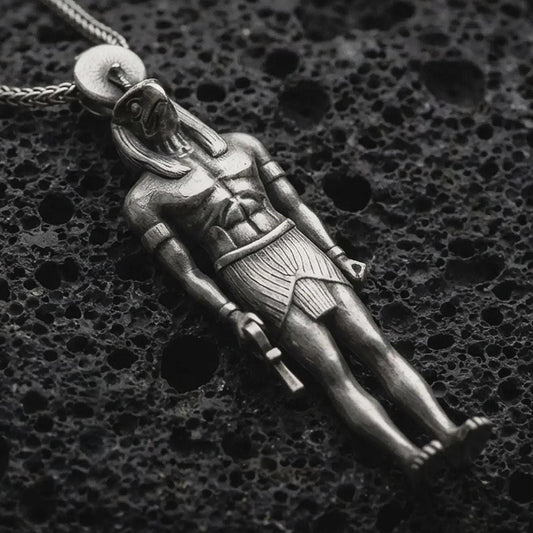 Oxidized Ra amulet pendant necklace resting on black volcanic stone, featuring an ancient Egyptian deity figure with a falcon-like head, sun disk, and strong sculpted body in a detailed macro product photograph.