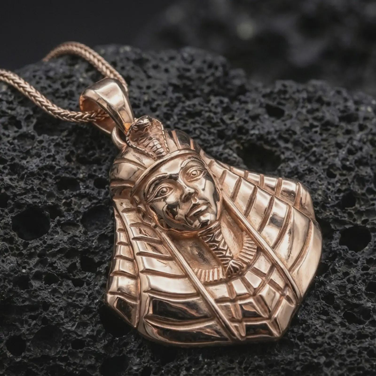 Rose gold plated Tutankhamun pendant necklace presented on textured volcanic stone, showcasing the pharaoh portrait, regal headdress, and refined antiquity-inspired design in a macro studio shot.