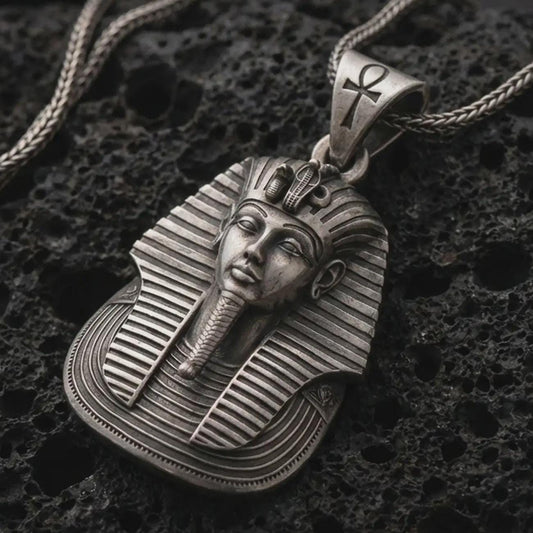 Oxidized Tutankhamun pharaoh pendant necklace resting on black volcanic stone, featuring a detailed Egyptian king face, striped nemes headdress, and ankh symbol on the bail in a macro product photograph.