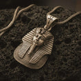 Load image into Gallery viewer, Bronze Tutankhamun pharaoh pendant necklace resting on black volcanic stone, featuring a detailed Egyptian king face, striped nemes headdress, and ankh symbol on the bail in a macro product photograph.
