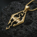 Load image into Gallery viewer, Gold plated Skyrim dragon pendant necklace on textured volcanic rock, premium close-up studio composition emphasizing the dragon symbol, geometric frame, and fantasy game-inspired design.
