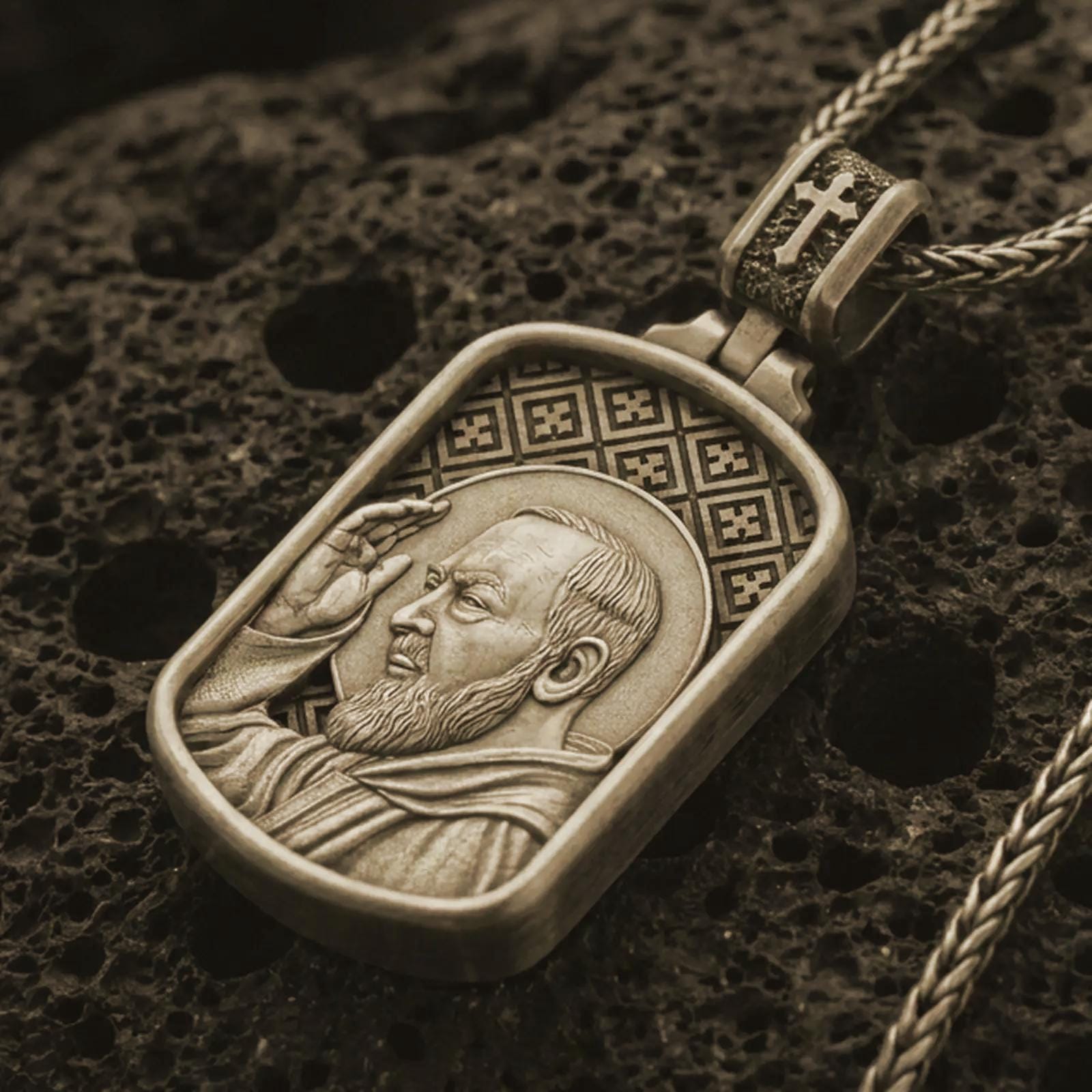 Bronze Saint Padre Pio dog tag pendant necklace on textured volcanic rock, close-up studio shot highlighting the saint profile, blessing gesture, halo, and antique silver contrast.