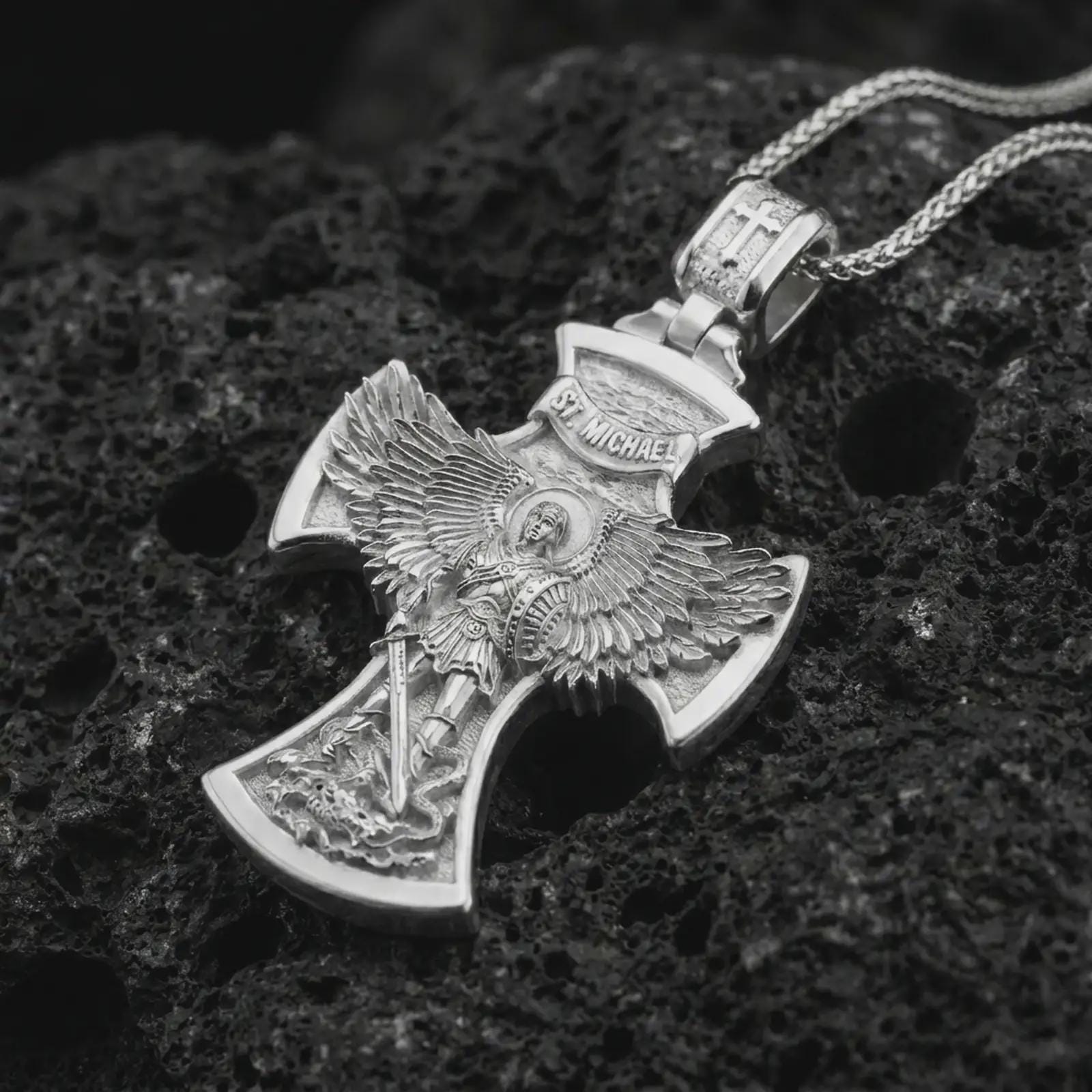 Polished silver Saint Michael cross pendant necklace on textured volcanic rock, centered macro jewelry photo with detailed archangel relief and bright silver highlights.