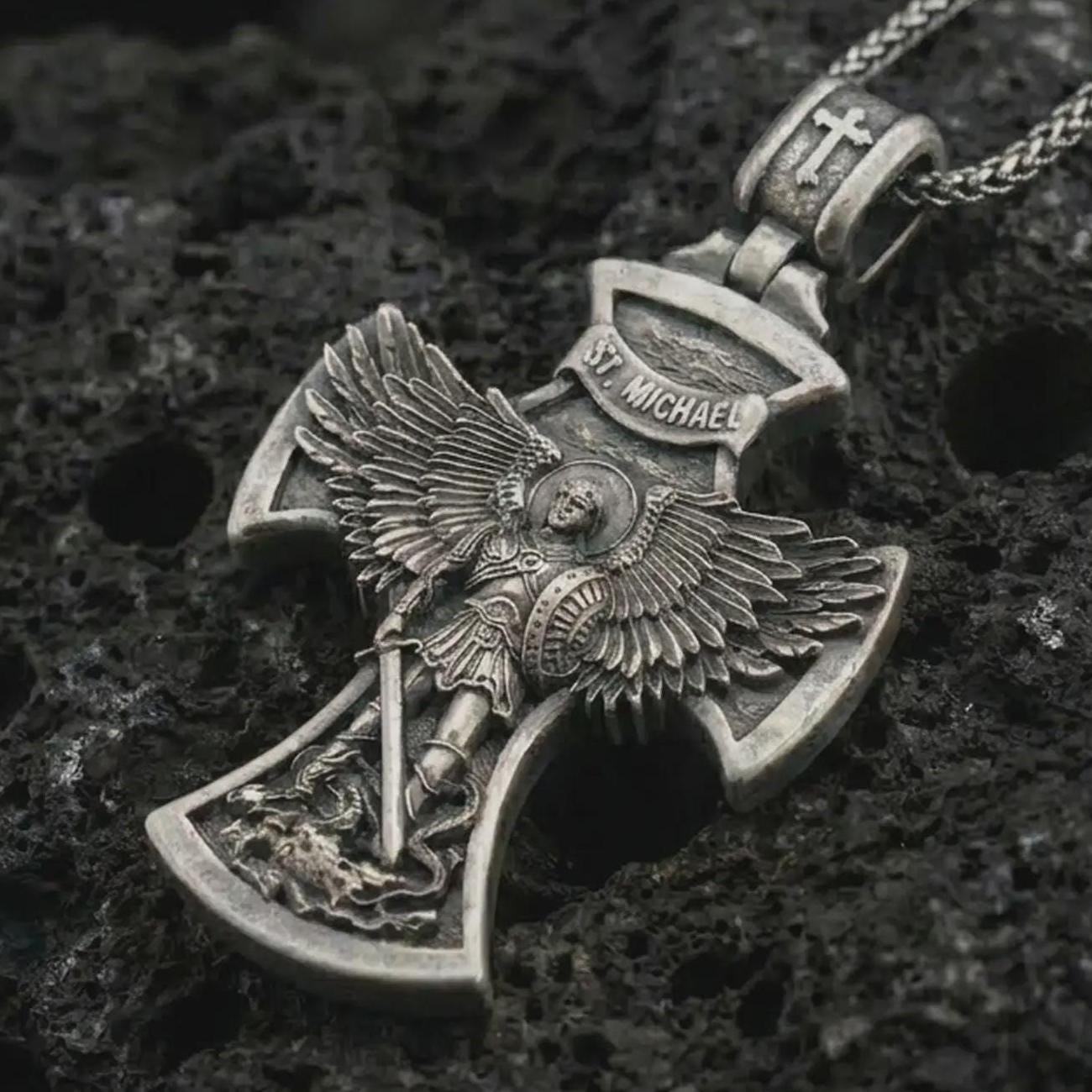 Oxidized Saint Michael cross pendant necklace on textured volcanic rock, close-up studio shot highlighting the archangel wings, sword, shield, demon detail, and antique silver contrast.