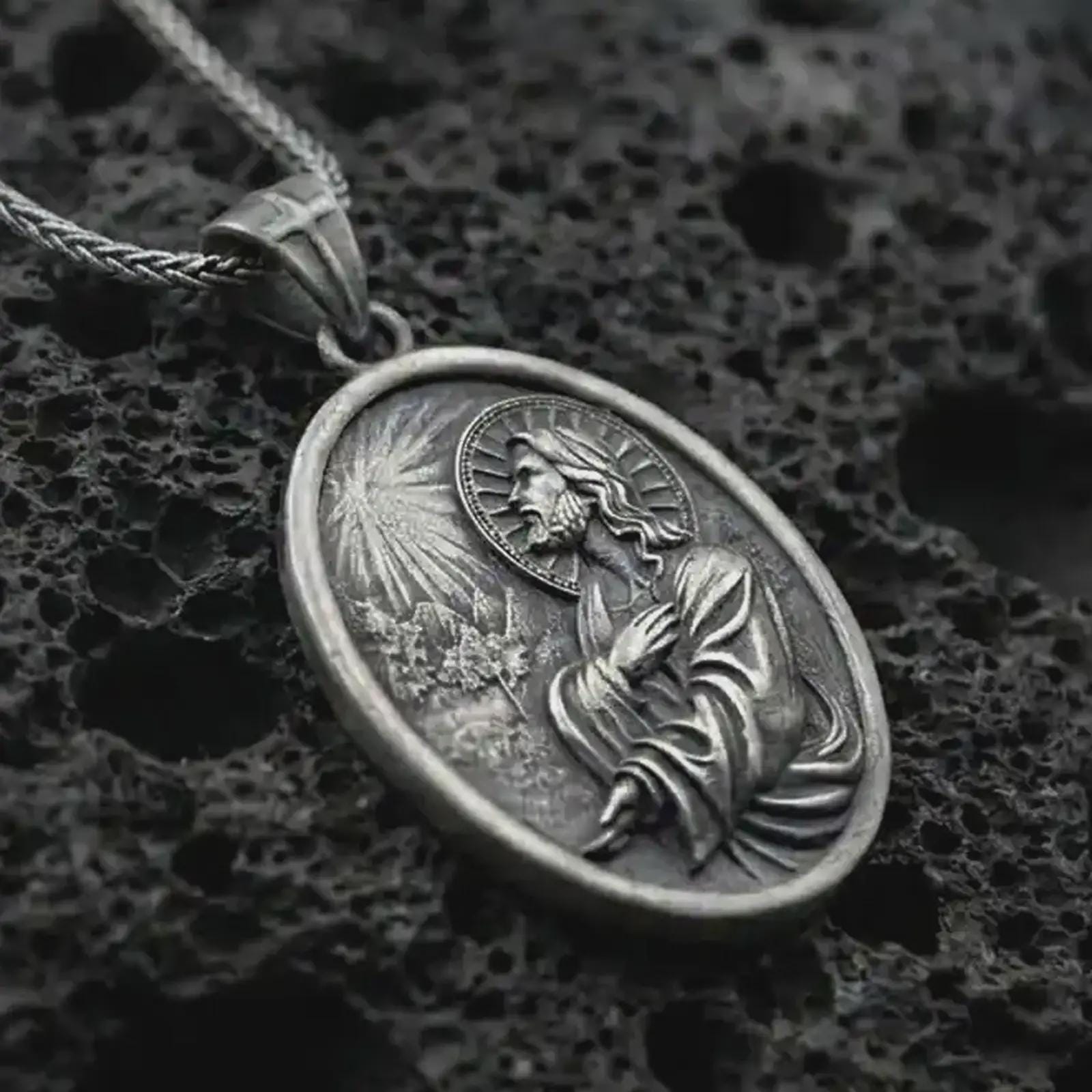 Oxidized Jesus Christ prayer pendant necklace on textured volcanic rock, close-up studio shot highlighting the praying Christ relief, radiant light detail, halo, and antique silver contrast.